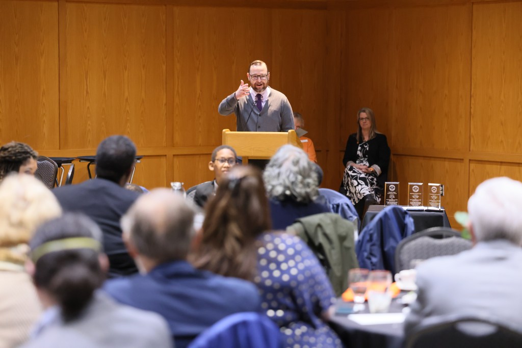 An image of K. Chad Clay giving a speech at a lectern next to a table holding glass awards. Two people sit behind him and the backs of the crowd in front of him can be seen.